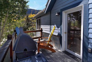 A wooden deck with a barbecue grill and a rocking chair, a door with a window, and a view of trees and hills. - The Red Lady, Cute 2BD in Town! (Crested Butte)
