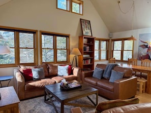 A cozy living room with leather sofas, a wooden coffee table, and a bookshelf. Large windows offer views of trees. - The Red Lady, Cute 2BD in Town! (Crested Butte)