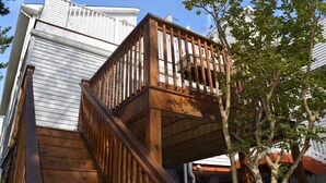 Un escalier en bois mène à une terrasse dotée d’une rambarde, qui est entourée d’arbres et bordée par un bâtiment blanc.