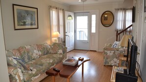 Un salon avec un canapé à motifs floraux, une table basse en bois, un téléviseur posé sur un meuble et une porte donnant sur un balcon.