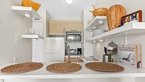 Cozinha moderna com armários brancos, micro-ondas, torradeira e cafeteira. Há jogos americanos trançados na bancada, e um livro intitulado 'The New Gleaners Sailing Manual' está visível.