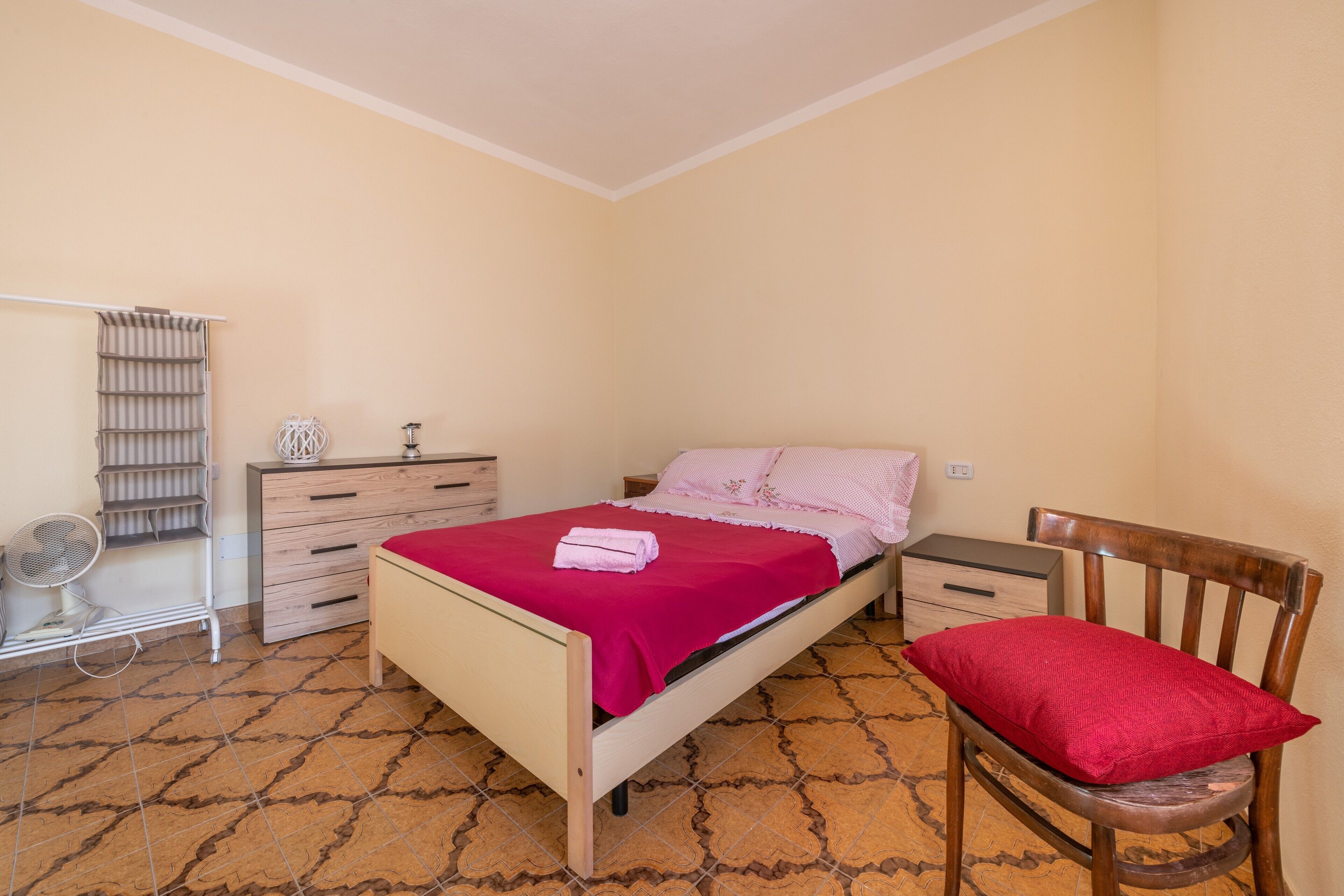 Un dormitorio con cama, una silla de madera, una mesita de noche, un aparador, un ventilador y una ventana.