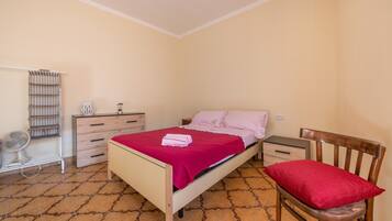 Un dormitorio con cama, una silla de madera, una mesita de noche, un aparador, un ventilador y una ventana.