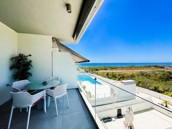 A balcony with a pool, white chairs, a table, and a potted plant.