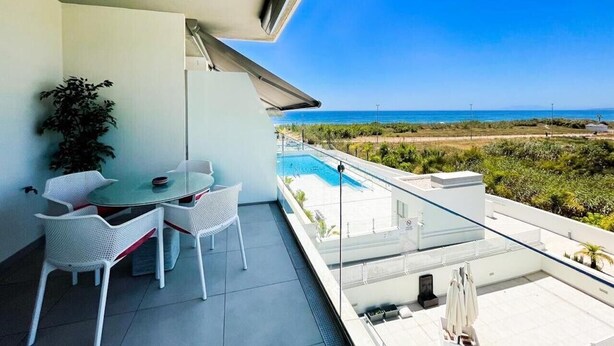 Un balcón con mesa y sillas, barandilla de vidrio y vistas a una piscina y una playa.