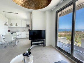 A modern living room with a TV, a dining area with chairs, a kitchen with cabinets, and a large window with a view of the ocean.