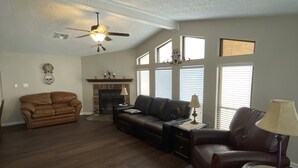 A spacious living room with hardwood flooring, a ceiling fan, a fireplace, and large windows with blinds. - Desert Hideaway (Glendale)