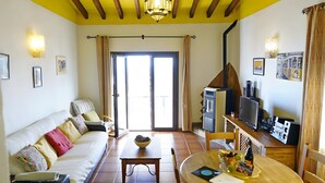 Una stanza con soffitto giallo, una ventola a soffitto, un divano, un tavolo da pranzo con sedie, un angolo cottura e una porta scorrevole in vetro che dà su un’area esterna.