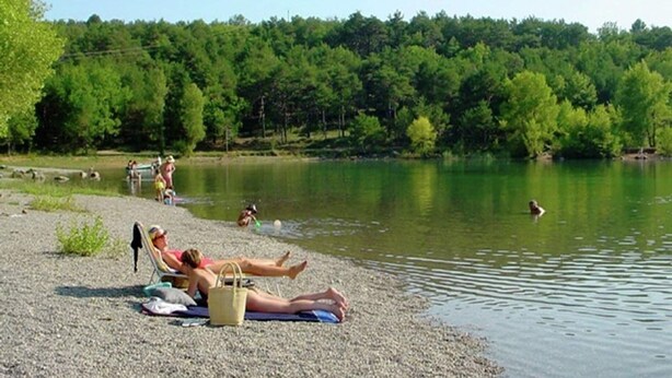 A lakeside setting with people relaxing on the shore, a clear view of the water, and a forested background.