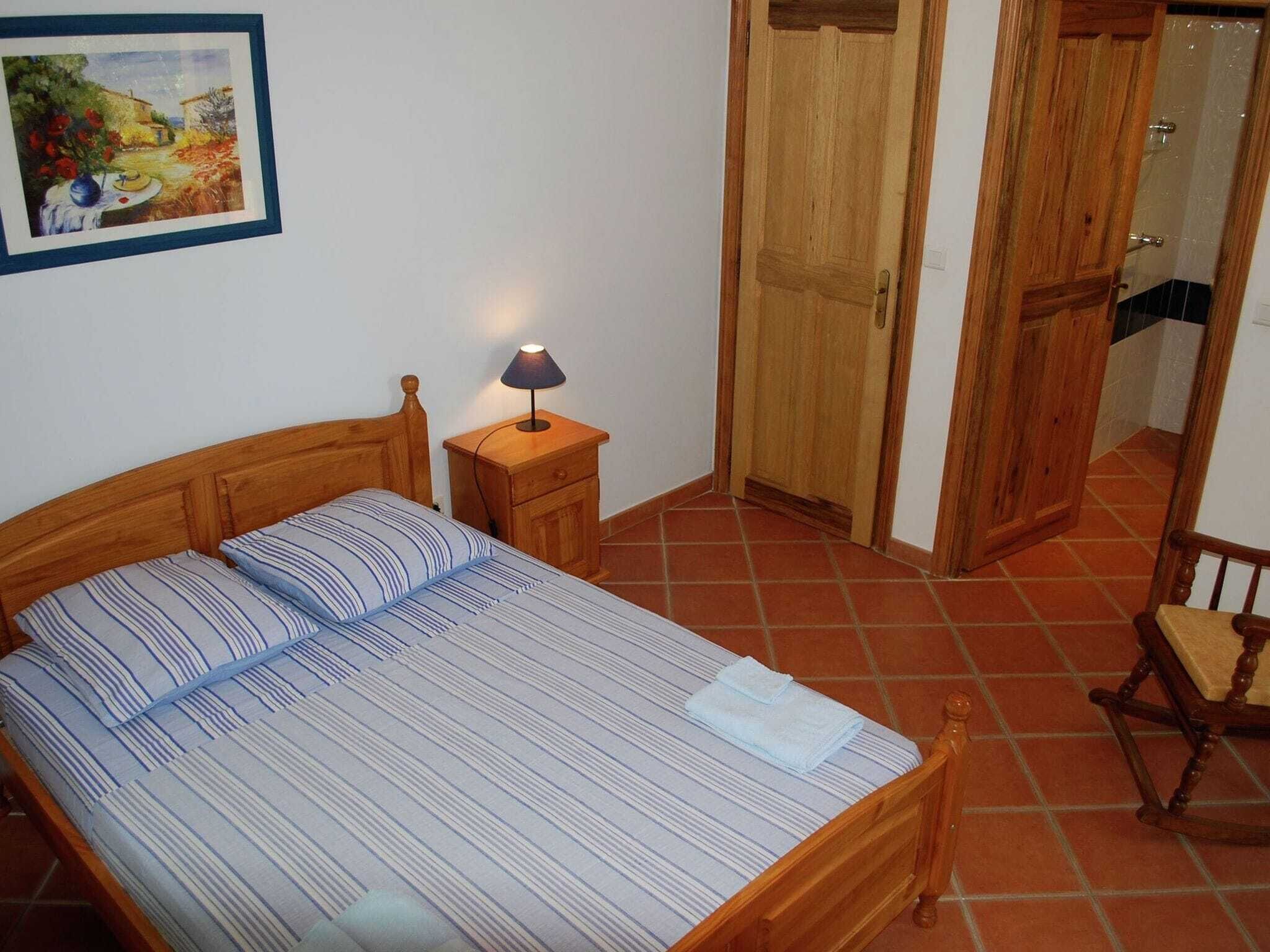 A bedroom with a wooden bed, a nightstand with a lamp, a wooden wardrobe, and a chair.