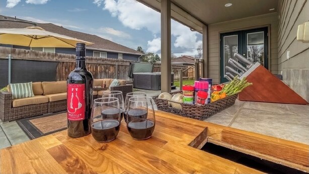 Une terrasse avec une table en bois, une bouteille de vin rouge et des verres, un panier en osier rempli de légumes, et un canapé en arrière-plan.