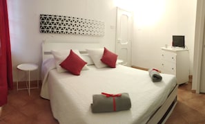 Una cama bien hecha con ropa blanca y almohadas rojas, una mesita de noche, un televisor montado en la pared y un armario con espejo.