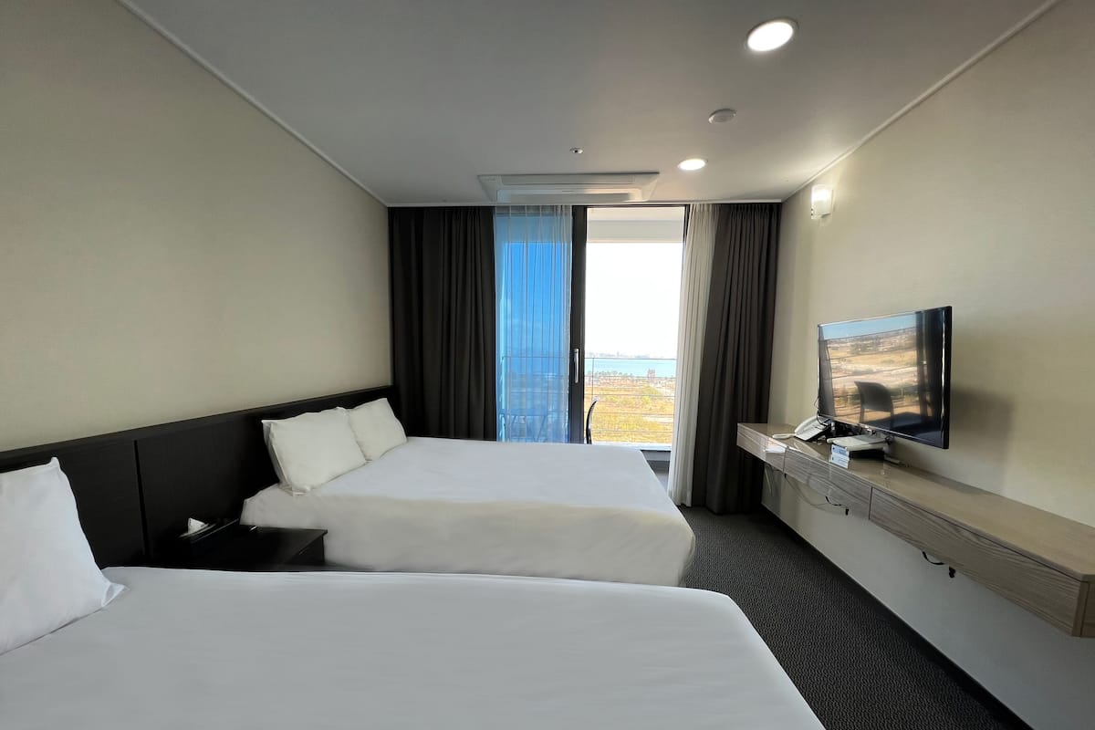 A hotel room with two beds, a desk with a flat-screen TV, and a large window offering a view of the outdoors.