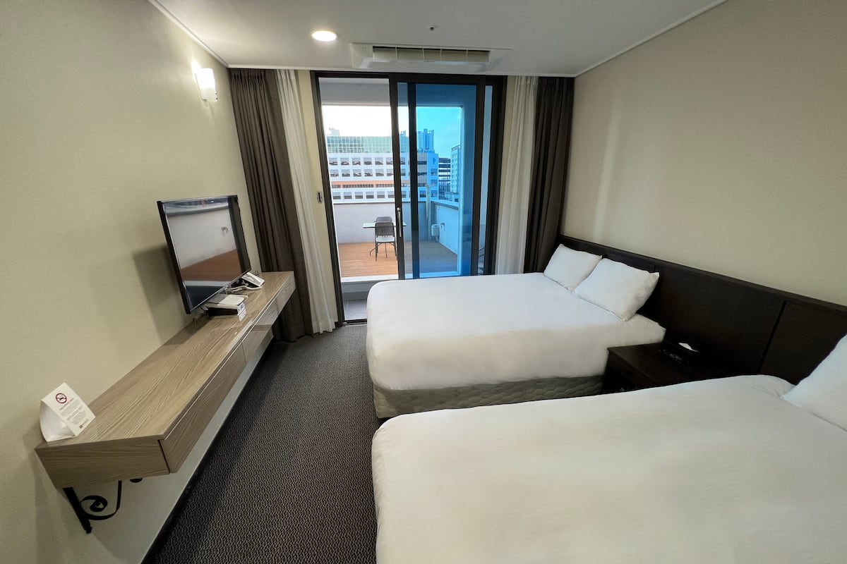 A hotel room with two beds, a TV, a desk, and a view of the city through the window.