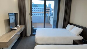 Hotellihuone, jossa on sänky, työpöytä, jossa on tietokone ja puhelin, ikkuna, josta on näkymä rakennuksiin, sekä liukuovi uloskäyntiä varten parvekkeelle.