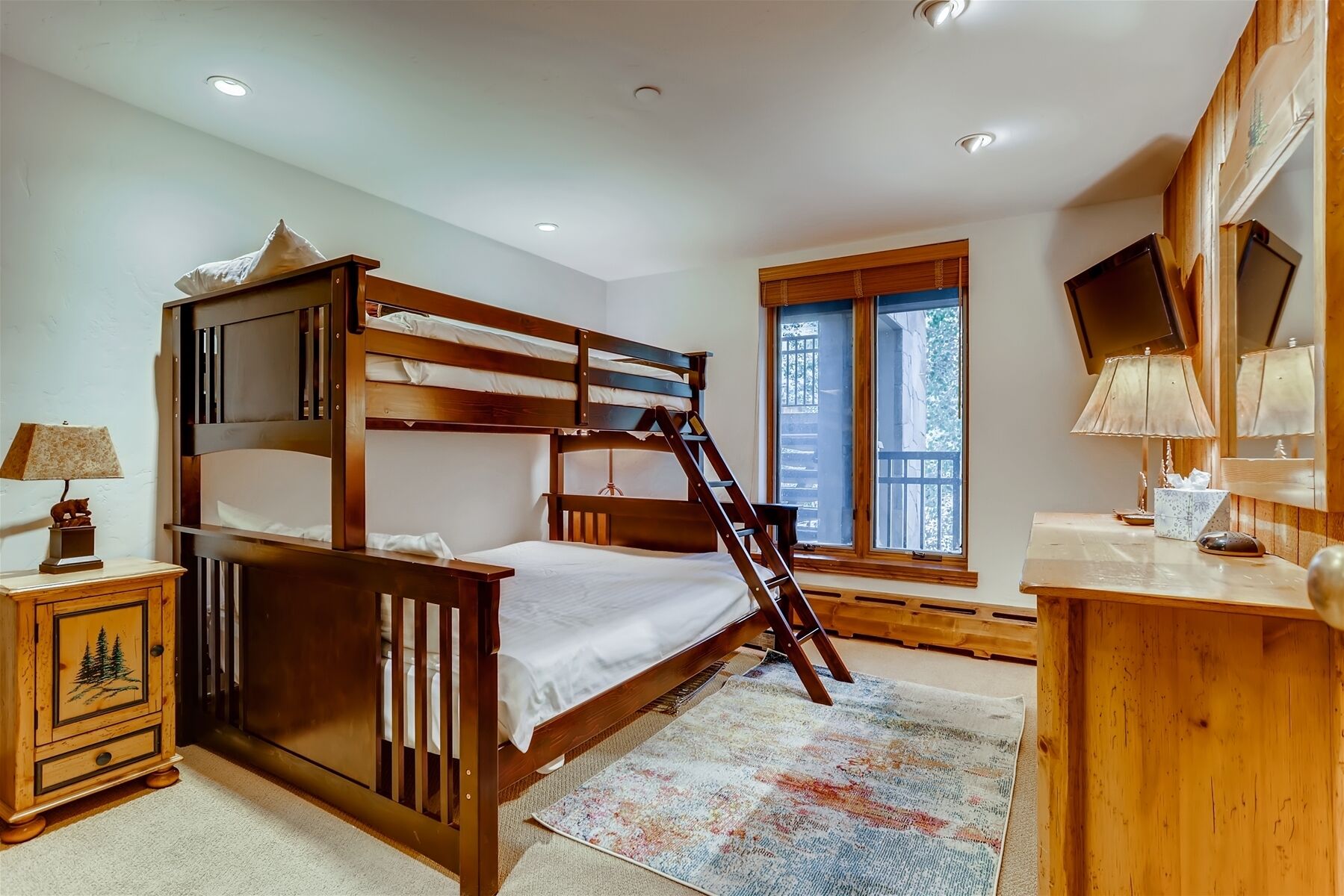 A bunk bed with a ladder, a desk with a lamp, a small cabinet with a tree design, and a window with a view of trees.