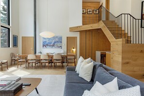 A spacious living area with a gray sofa, wooden dining table, and chairs. A staircase with a wooden railing leads to an upper level.