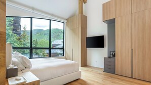 A modern bedroom with a large window, a bed, a bedside table, a flat-screen TV, and wooden cabinetry. - Tyrolean 6 | Newly Updated 4BR Condo Steps to Vail Village (Vail)