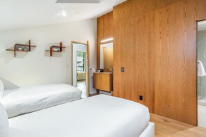 A modern bedroom with two beds, wooden wardrobe, and a bathroom visible through an open door.