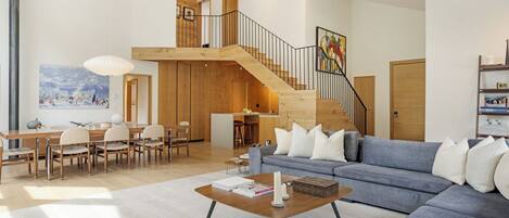 Amplia sala de estar con un sofá azul, una mesa de comedor de madera y una escalera que sube a un nivel superior.