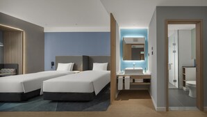 A modern hotel room with two beds, a desk with a sink, and a bathroom visible through an open door.