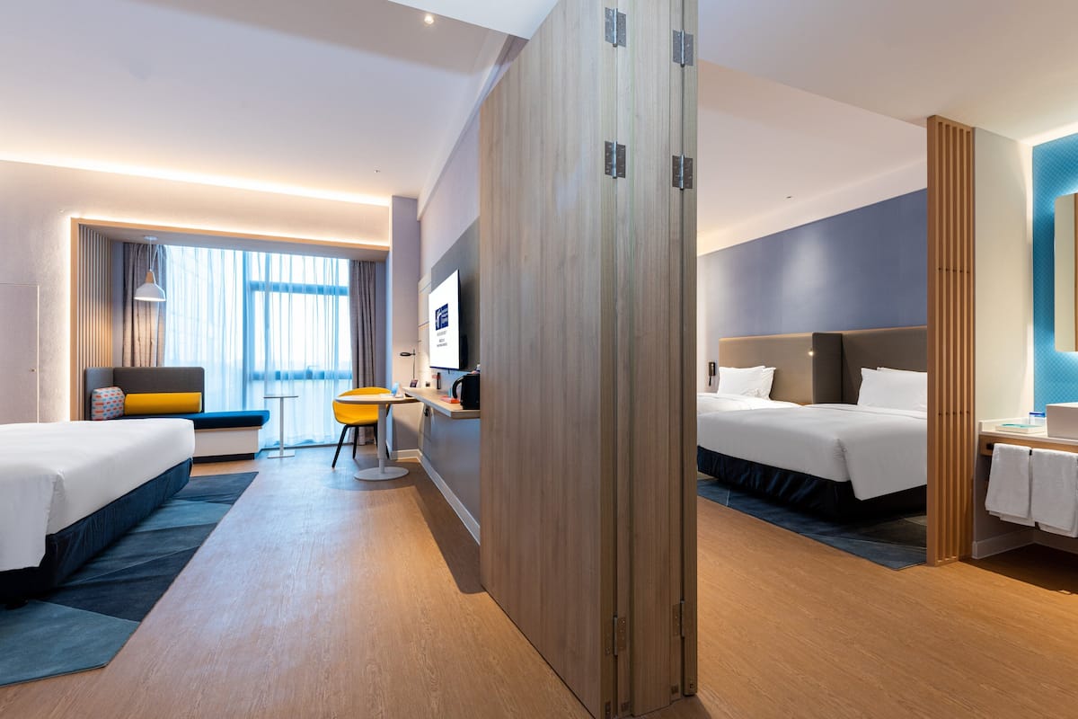 A modern hotel room with two beds, a desk, a chair, and a large window with curtains.
