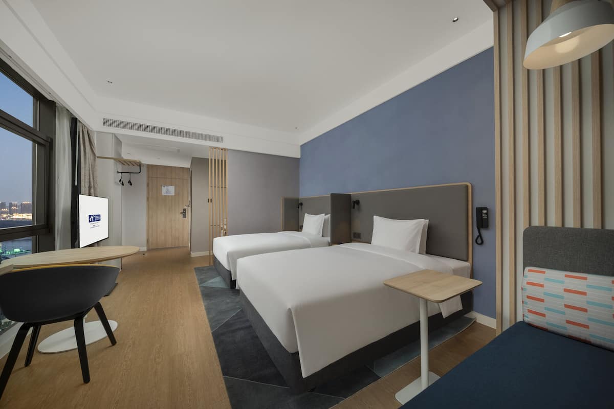 A hotel room with two beds, a desk, and a chair. There is a window with a view of the city.