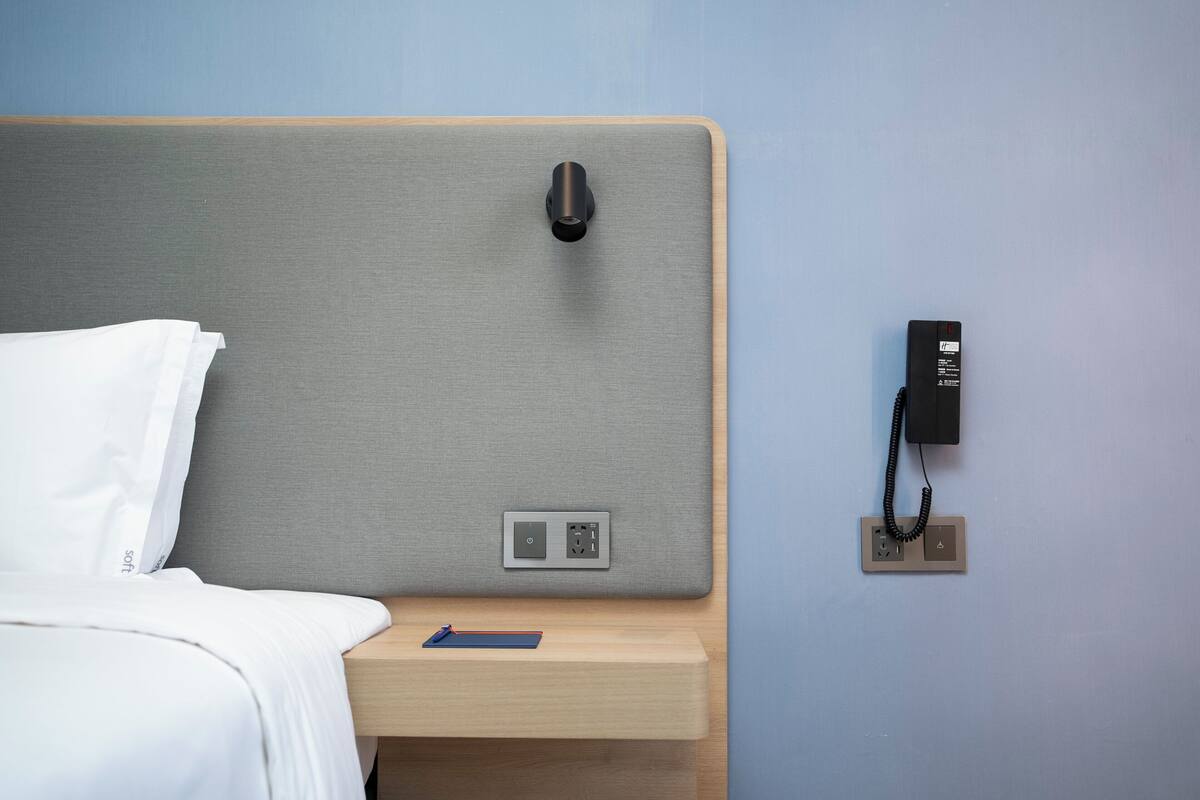A neatly made bed with white linens, a bedside table with a blue object, a power outlet, and a telephone on the wall.