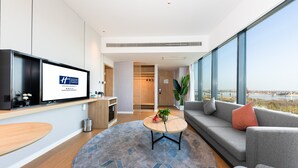 A modern hotel room with a large window, a round coffee table, a sofa, and a TV mounted on the wall.
