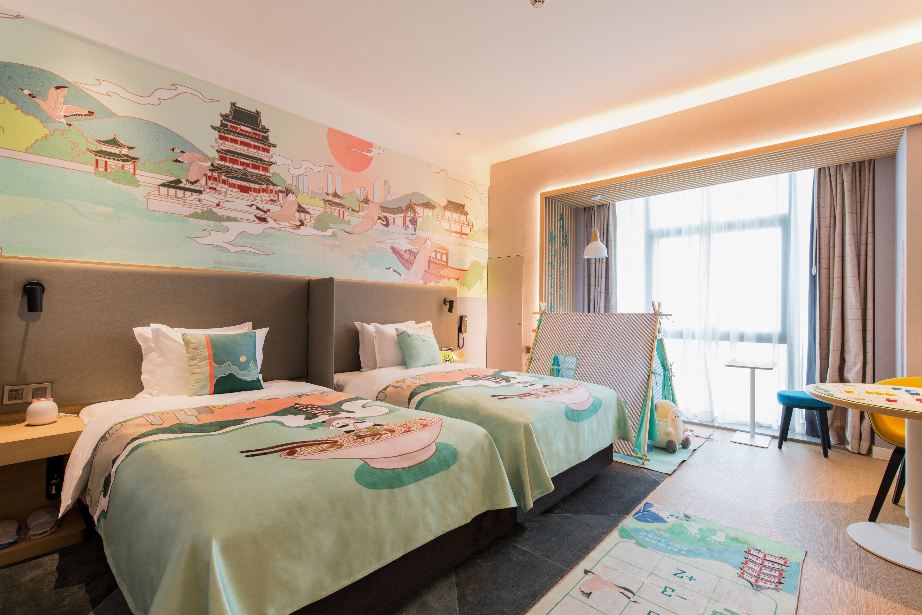 Habitación de hotel con dos camas, un mural grande en la pared, zona de comedor con mesa y sillas, y vistas al exterior.