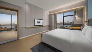 A modern hotel room with a large bed, a city view, a framed certificate on the wall, and a small seating area by the window.