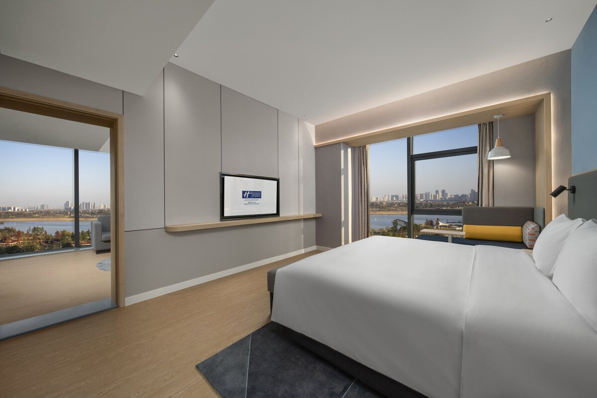 A modern hotel room with a large bed, a city view, a framed certificate on the wall, and a small seating area by the window.