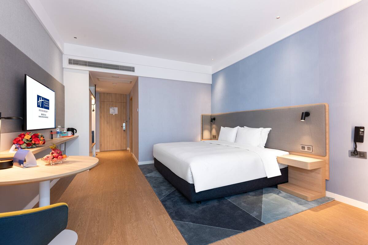 A modern hotel room with a large bed, a desk with a computer, and a TV on the wall.