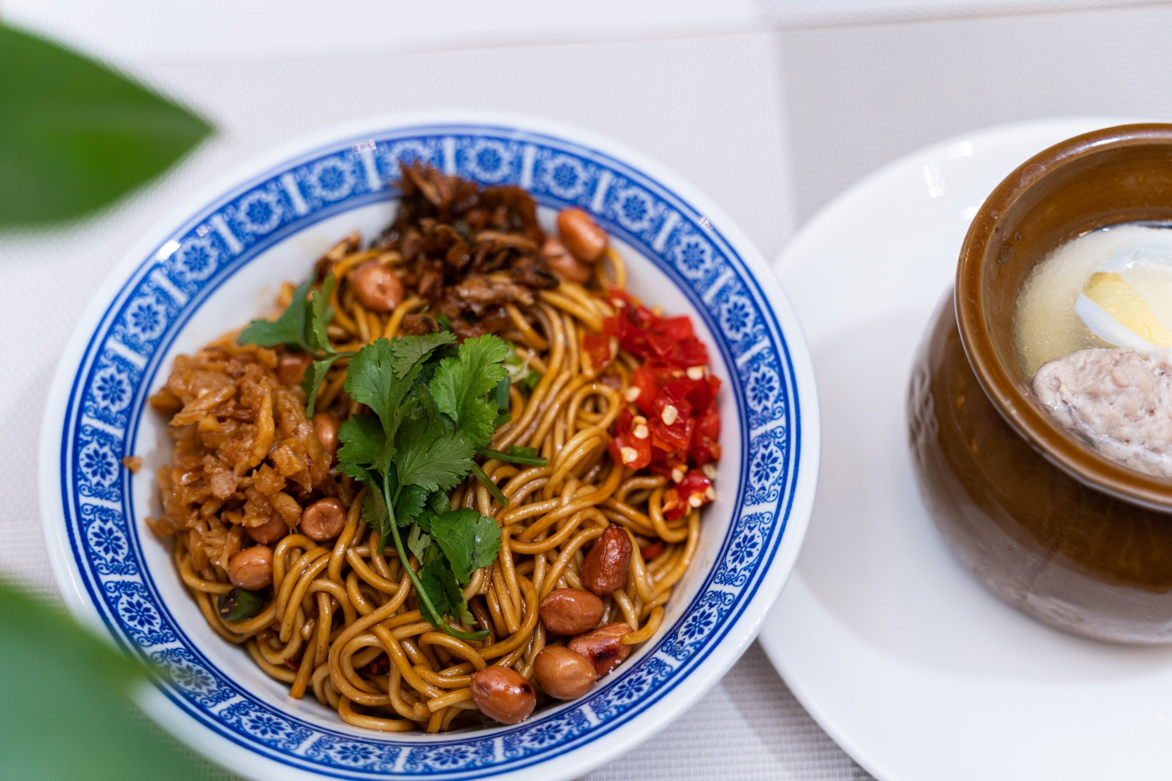 Un tazón de fideos con cacahuates y chiles rojos, adornado con cilantro, y un tazón de sopa con huevo duro y carne.