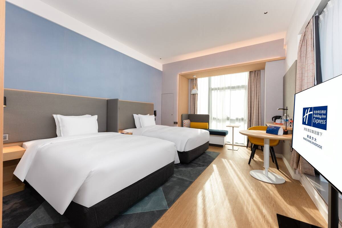 A hotel room with two beds, a desk, a chair, and a large window with curtains.
