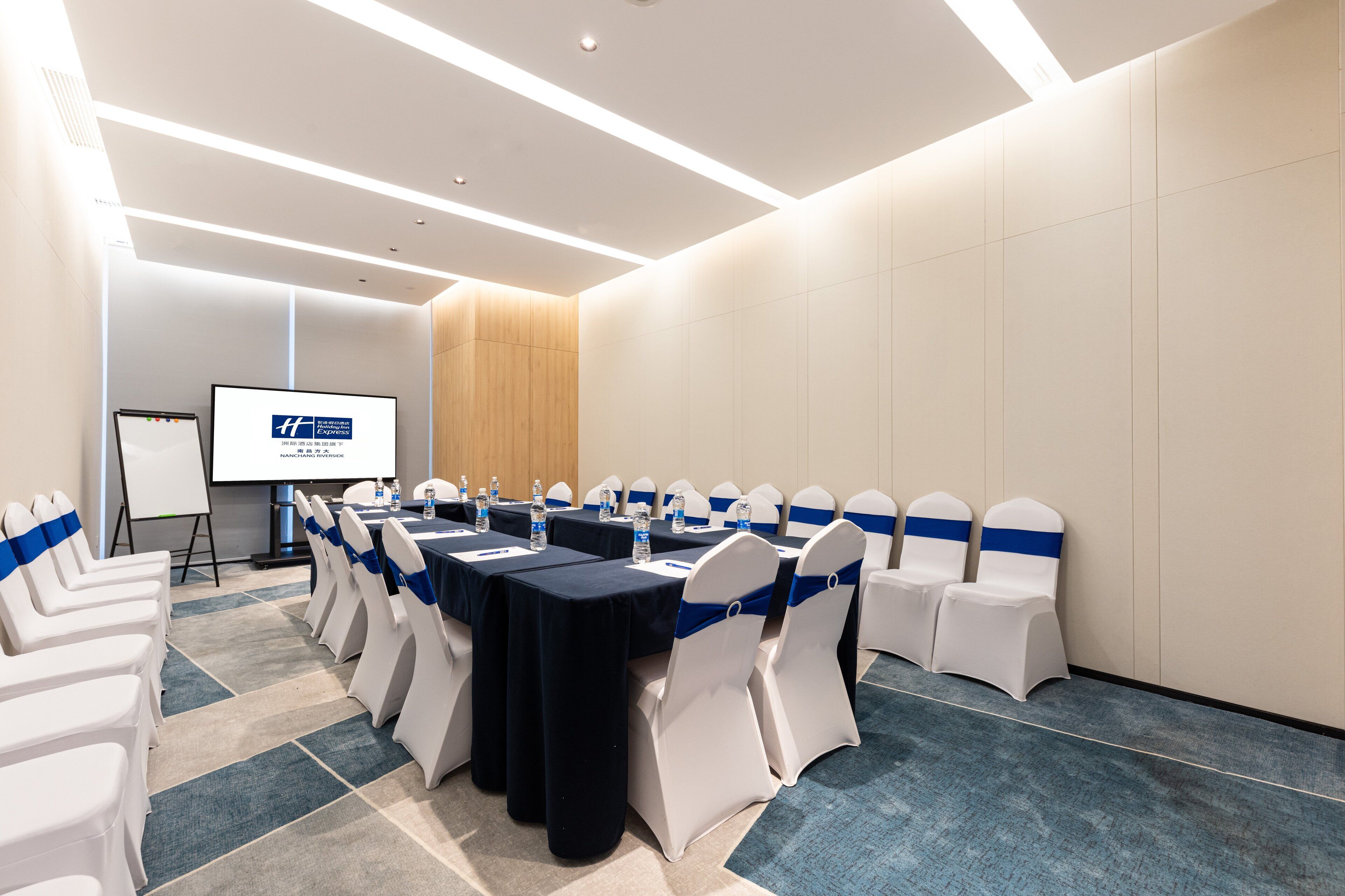 Una sala de conferencias con una larga mesa, sillas blancas con cintas azules, una pizarra y una pantalla para proyector.