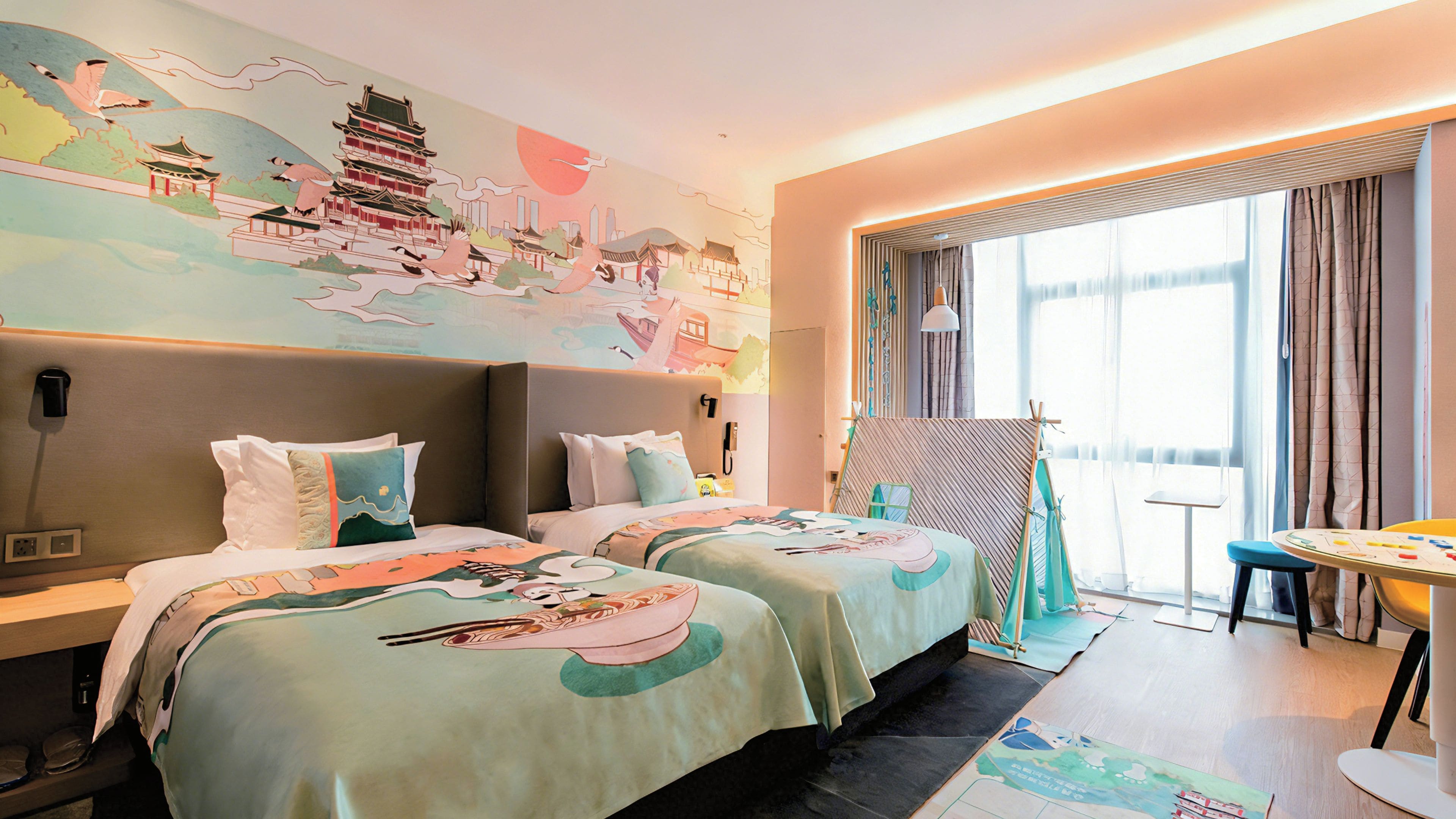 A hotel room with two beds, a large window with curtains, a desk, and a wall mural.