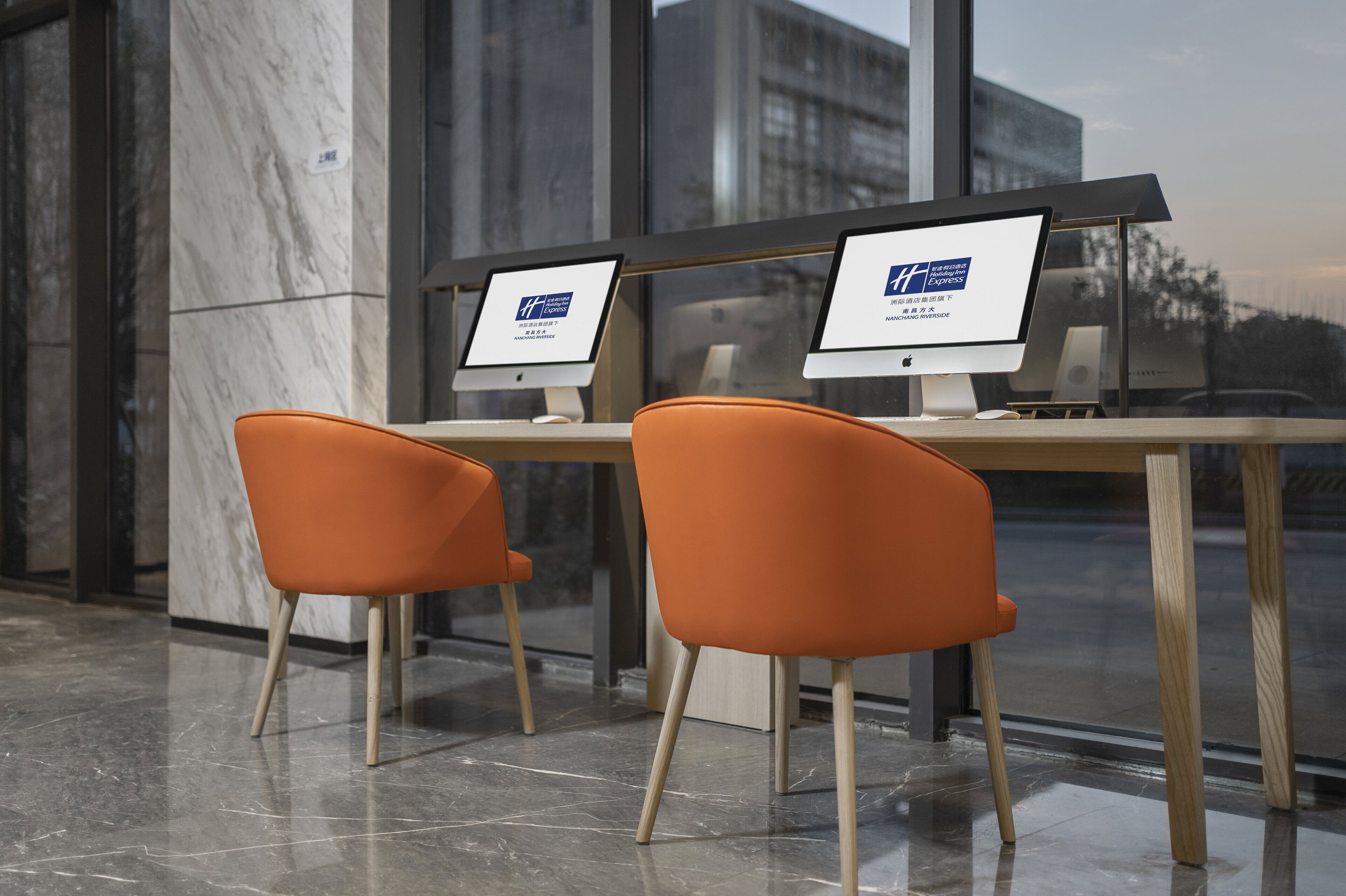 A modern hotel lobby with two orange chairs, a marble floor, and two Apple iMac computers on a wooden table.