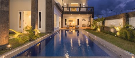 A modern house with a swimming pool, a balcony, and a well-lit interior.