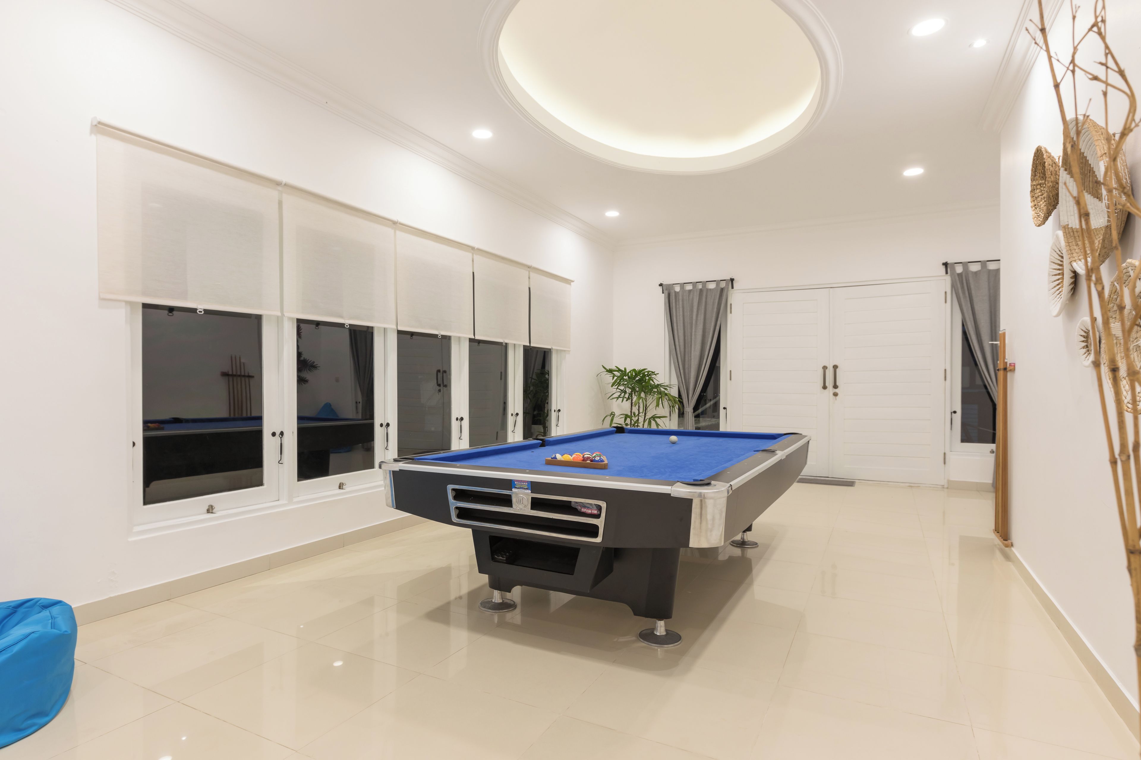 A modern indoor pool table with a blue felt surface, surrounded by white cabinets and a large window with blinds.