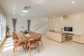 A modern living room with a wooden dining table, beige sofas, a TV, and a staircase.