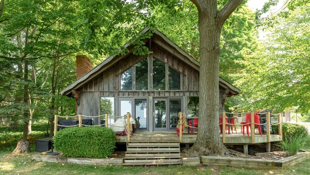 A rustic cabin with a wooden deck, a covered porch, and a large tree in front.