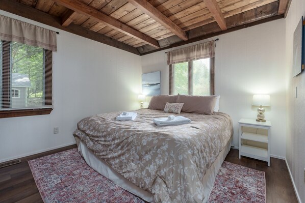 A bedroom with a large bed, two bedside tables, a wooden ceiling, and a window with blinds.