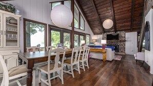 A spacious dining area with a wooden table and white chairs, a stone fireplace, and large windows offering views of the outdoors.
