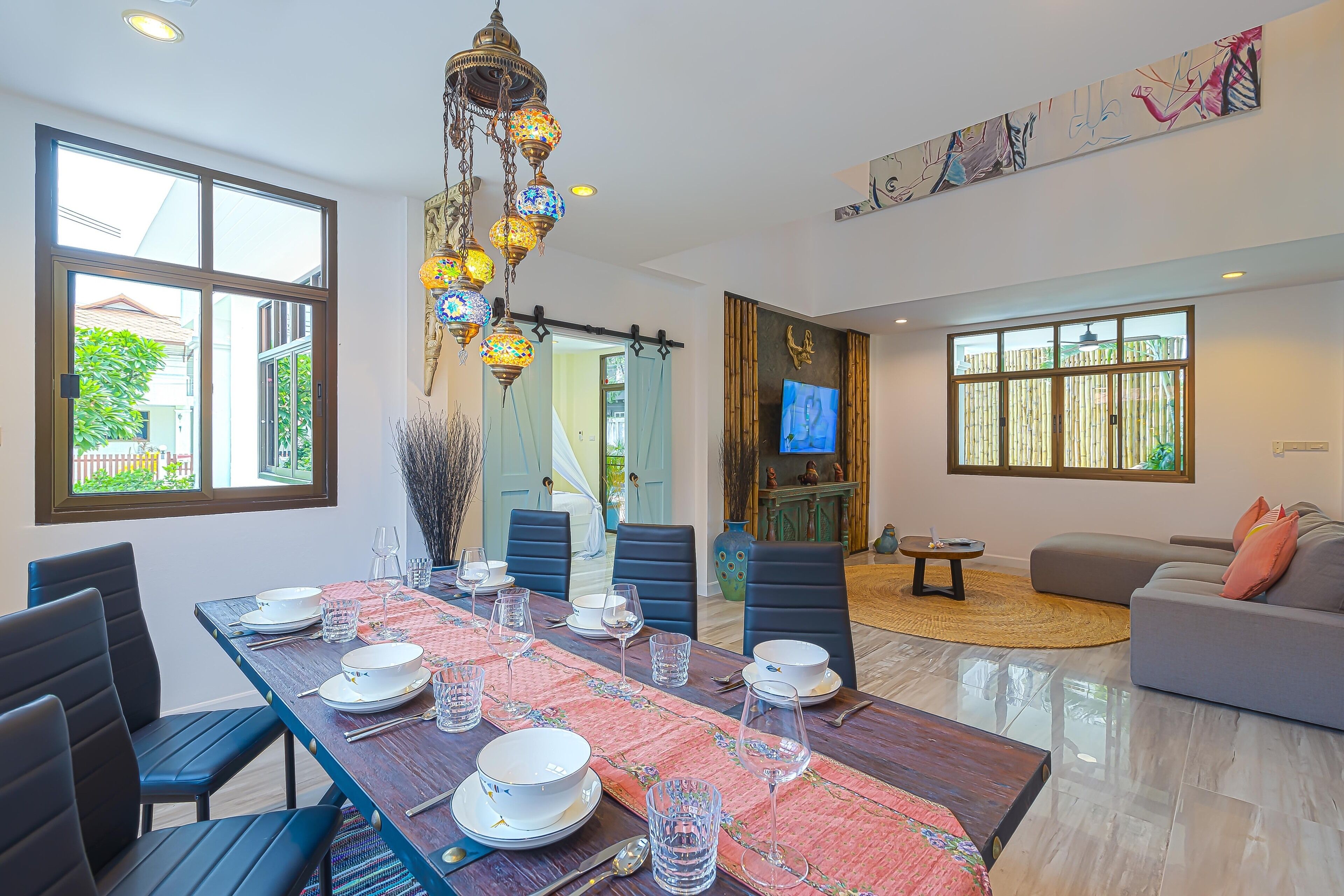 Comedor con una larga mesa dispuesta para seis personas, rodeada de sillas, una lámpara colgante decorativa y un amplio ventanal con vistas a zonas verdes.