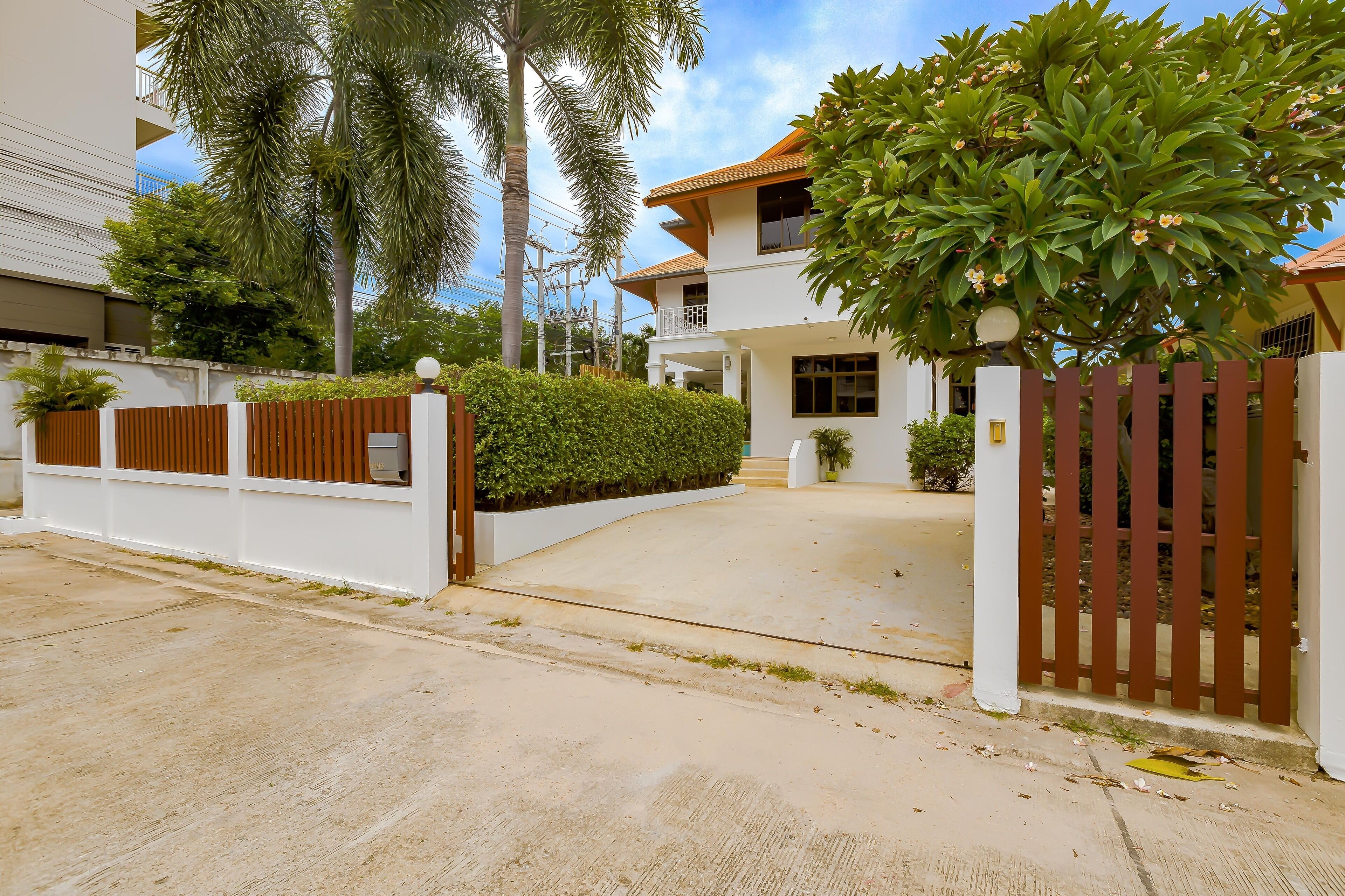 A modern two-story house with a gated entrance, surrounded by lush greenery and palm trees.