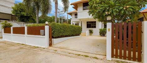A modern two-story house with a gated entrance, surrounded by lush greenery and palm trees.