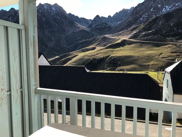 A mountain view from a balcony with a white railing, overlooking a valley with a dirt road and a small building.