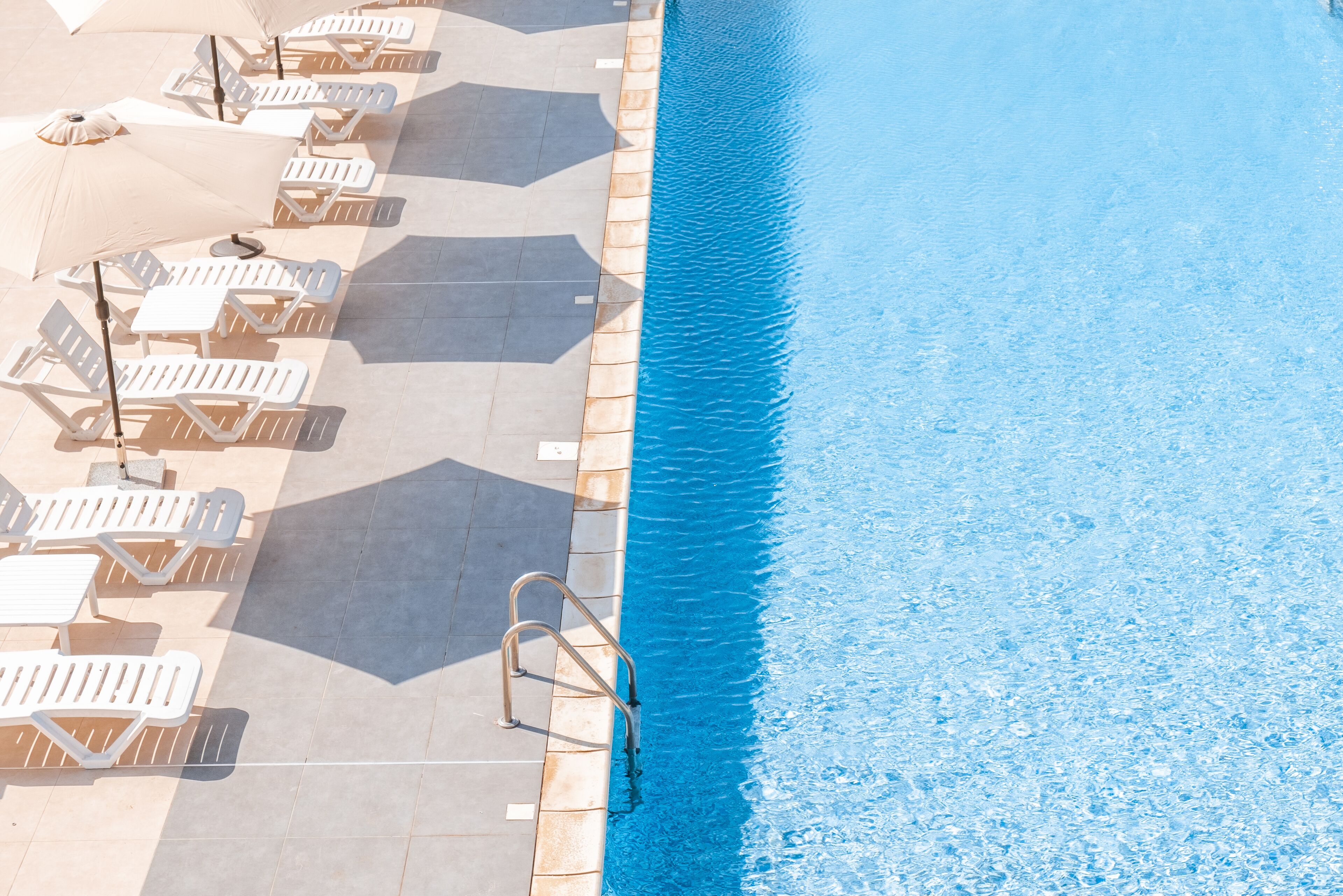 Zona de piscina con sillas de descanso blancas y sombrillas, una piscina azul cristalina y un camino empedrado.