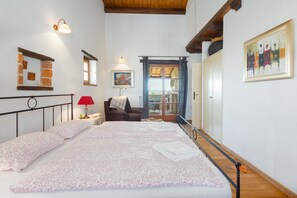 A bedroom with a large bed, a nightstand with a lamp, a chair, a window with curtains, and a door leading outside.